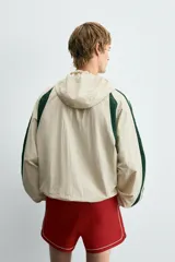 Cazadora de corte relajado confeccionada en tejido técnico ligero de color beige con bandas verdes a contraste en las mangas y hombros. Presenta capucha ajustable, cierre frontal con cremallera, forro interior de red y bolsillos laterales ocultos.