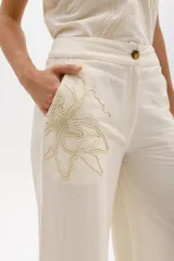 Pantalón de lino color crudo, de corte amplio y tiro alto con cierre frontal. Presenta un bordado artesanal en hilo beige con diseño floral abstracto en el lateral del bolsillo.