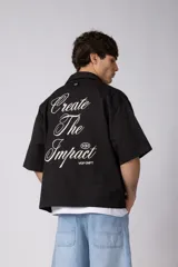 Camisa de manga corta negra, de corte boxy, con cuello camisero, cierre frontal de botones y bordado circular en el pecho.