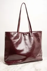 Bolso tipo tote de gran tamaño en color bordó con acabado brillante. Cuenta con doble asa de hombro y placa metálica decorativa con logo en la parte frontal.