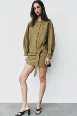 Vestido corto verde militar de cuello solapa y manga larga, con bolsillos de plastrón en el delantero y cierre frontal con cremallera oculta por sopata. Cuenta con un cinturón incorporado del mismo tejido con hebilla metálica y aberturas laterales en el bajo.