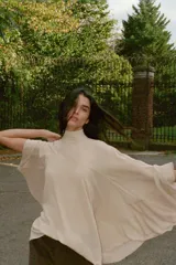 Top tipo capa de color beige, con cuello subido y cierre posterior con botones ocultos. Presenta un diseño fluido con bajo asimétrico y mangas acampanadas.