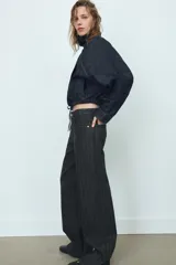 Jeans de tiro bajo y corte recto, confeccionados en denim negro con estampado de rayas verticales finas. Presentan cinco bolsillos, trabillas para cinturón y cierre frontal con cremallera y botón metálico.