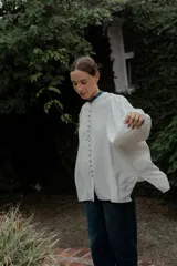 Camisa de corte oversize color crudo, con cuello mao y puños con detalle de volados en color azul. Presenta cierre frontal con botones forrados y mangas largas abullonadas.