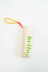 Paraguas plegable de color beige con la palabra 'hello.' estampada en color verde. Incluye una correa de muñeca de color amarillo con detalles en fucsia.