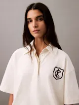 Chomba color crema de punto de algodón con cuello clásico, tapeta de botones oculta y mangas cortas. Presenta un corte holgado y un escudo bordado con el logo de Calvin Klein en el pecho.