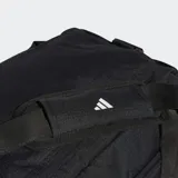 Bolso deportivo tipo duffel Adidas Essentials, color negro, con tres franjas blancas y logo de la marca en el lateral. Presenta compartimento principal amplio, bolsillos frontales y laterales con cierre, y correa de hombro ajustable.