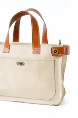 Bolso tipo tote color beige con detalles en cuero marrón. Tiene doble asa corta y correa larga desmontable. Cuenta con bolsillo externo y cierre con botón metálico. Mide 30 x 10 x 21 cm.