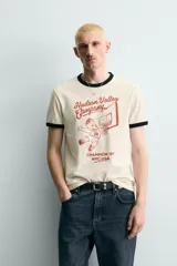 Camiseta de corte regular fit, color crema, con cuello redondo y mangas cortas. Presenta un estampado frontal con un personaje de dibujos animados jugando al baloncesto y texto "Hudson Valley Company Champion '87 NYC, USA". Los acabados del cuello y las mangas son de vivo negro a contraste.