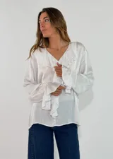 Blusa de satén color blanco con escote en V, cuello con volados y mangas largas con puños también decorados con volados.