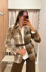 Blazer de largo medio con diseño a cuadros en tonos tierra, confeccionado en mezcla de algodón y poliéster. Presenta solapas clásicas, bolsillos laterales y un cinto de cuero fino incorporado para ajustar la cintura.