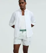 Bermuda de baño para hombre confeccionada en microfibra ligera, color blanco, con una estampa sutil de cocoteros en tonos verdes/azulados. Presenta cintura elástica con cordón de ajuste contrastante en color verde, y bolsillos laterales.