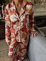 Pijama de seda estampado con diseño botánico en tonos rojizos y naranjas, compuesto por una camisa de manga larga con solapas, botones frontales y bolsillo en el pecho, y pantalón recto a juego.