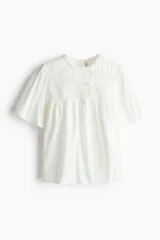 Blusa blanca de algodón con cuello redondo ribeteado en encaje y mangas cortas holgadas. Presenta un canesú fruncido con jaretas y detalles de broderie en la parte frontal.