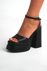 Sandalias negras de cuero sintético, con plataforma y taco cuadrado. Tira delantera ancha y pulsera al tobillo con hebilla.