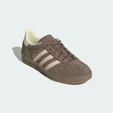 Championes Adidas Gazelle Indoor de diseño clásico, confeccionados en gamuza color marrón con las icónicas tres tiras laterales y refuerzo en el talón en color rosado. Presentan suela de goma translúcida y cierre de cordones.