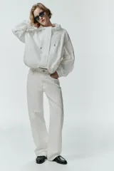 Chaqueta blanca de cuello redondo con capucha ajustable mediante cordones elásticos. Presenta manga larga con puños elásticos, bolsillos delanteros y bajo asimétrico ajustable con cordones y stoppers. El cierre frontal combina cremallera y botones ocultos bajo una solapa.