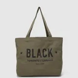Bolso tipo tote bag de lona color verde militar, con estampado frontal que incluye texto en negro y estrellas a los lados. Cuenta con dos asas superiores para llevar al hombro.