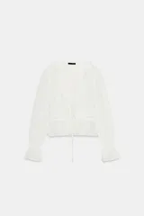 Camisa de gasa color blanco con escote en pico, cierre frontal con lazadas y detalles de volantes. Presenta mangas largas con puños elásticos y terminación en volante, además de bordados a tono.