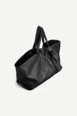 Bolso tipo shopper de cuero negro con textura granulada. Presenta doble asa de mano, cierre magnético y un detalle decorativo de nudo con argolla metálica en el frente.