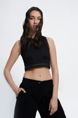 Top corto negro de cuello redondo y corte tipo halter, confeccionado en gabardina de algodón. Presenta costuras a la vista en color blanco, pinzas delanteras para mayor ajuste y cierre lateral.