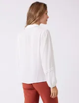 Camisa de gasa blanca con cuello solapa y cierre frontal de botones. Presenta un delicado galón de encaje festoneado a lo largo de la cartera frontal. Sus mangas largas son ligeramente abullonadas, con insertos de puntilla horizontal y puños elásticos fruncidos.