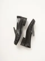 Mocasines de cuero negro con costuras pespunteadas en hilo blanco a contraste, diseño fruncido en la puntera, talón plano con pequeño tacón y apliques sutiles en charol.