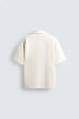 Camisa de manga corta con cuello bowling y calce relaxed fit, confeccionada en tejido seersucker elástico con textura. Presenta un diseño a rayas verticales en tonos oscuros y cierre frontal con botones.