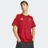 Camiseta de fútbol Adidas de la primera equipación de Bélgica, color rojo con patrón sutil de llamas y demonios. Presenta cuello redondo con ribete negro y amarillo, y detalles de las tres franjas amarillas en los hombros. El escudo de la RBFA está bordado en el pecho.