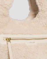 Bolso tote de sherpa color crema, con asas de mano y correa bandolera ajustable. Cuenta con cierre de cremallera y bolsillos internos.