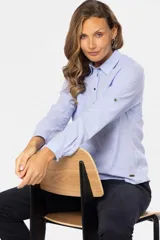 Camisa blanca de manga larga con cuello y tapeta de medio botón. Presenta dos bolsillos frontales con tapa y botones metálicos. Las mangas tienen presillas para sujetar el puño enrollado.