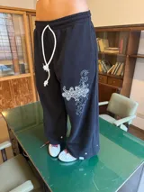 Pantalón de jogging negro de corte baggy, confeccionado en algodón. Presenta una estampa plastisol con bordado decorativo en la pierna izquierda, cintura elástica con cordón largo ajustable y botones en el ruedo para regular la botamanga.