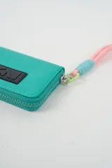 Billetera pequeña color turquesa con cierre perimetral y parche frontal negro con logo. Incluye una correa de muñeca de cordón rosado con detalle de hilo celeste.