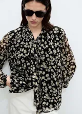 Blusa de gasa con estampado floral de margaritas sobre fondo negro, diseño recto semitransparente con cuello pico y detalle de lazo frontal. Cuenta con mangas largas y forro interior.