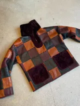 Buzo de cuero genuino con diseño de patchwork en cuadros de colores negro, marrón y bordó. Presenta cuello alto y bolsillos frontales con detalle de piel sintética en color bordó.