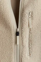 Campera deportiva de tejido sherpa color beige, con cuello elevado, cierre frontal completo, mangas raglán, bolsillo superior con cierre y bolsillos laterales. Presenta terminaciones elásticas en puños y ruedo.