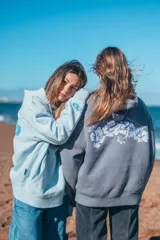 Hoodie gris con capucha y cordones ajustables, de corte holgado y manga larga. Presenta un bordado de flores blancas en la espalda y en el puño.