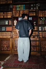 Camiseta de estilo deportivo con cuello polo, confeccionada en tejido técnico negro con finas rayas verticales blancas. Presenta paneles laterales en gris, mangas cortas con bordes en contraste y detalles gráficos estampados en el pecho.