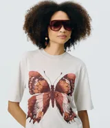 Remera oversized de algodón color gris claro, con cuello redondo y mangas cortas. Presenta una estampa frontal de mariposa marmolada en tonos tierra con pequeños detalles de strass.