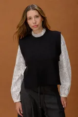 Chaleco de punto negro con cuello redondo y corte recto. Presenta aberturas laterales amplias con detalle de tira y botón, ofreciendo un diseño versátil y moderno para superposiciones.