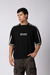 Remera de tejido de punto con textura acanalada, color negro, de corte relajado y manga corta. Presenta cuello en V, detalles de franjas blancas en los hombros y mangas, y un estampado gráfico central en color blanco.