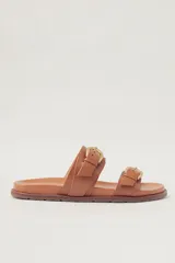 Sandalias planas de cuero color tostado, con dos tiras anchas ajustables mediante hebillas metálicas doradas. Diseño minimalista con suela flexible.
