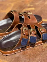 Sandalias de cuero marrón con tres tiras ajustables con hebillas doradas y detalles en rojo.