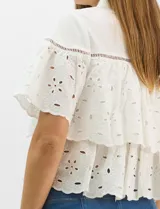 Camisa de algodón blanca con cuello clásico y cierre frontal de botones. Presenta un diseño con capas superpuestas y volados, combinando paneles de tela lisa con tejido bordado suizo de motivos calados y una cenefa decorativa. Posee mangas cortas y una silueta acampanada.