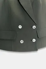 Chaleco de vestir color verde militar, estilo cruzado con doble botonadura plateada. Presenta cuello tipo solapa y diseño halter que deja la espalda descubierta. Cuenta con falsos bolsillos con tapetas en el delantero.