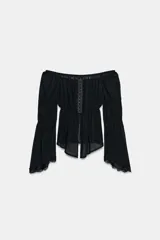 Top negro de manga larga con escote recto estilo off-shoulder, cierre frontal con ganchos metálicos y bajo asimétrico. Las mangas presentan frunces y terminan en puños acampanados con detalle de encaje.