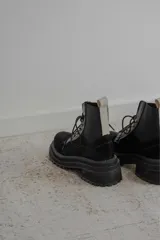 Borcego de gamuza color chocolate con paneles laterales elásticos negros y suela track dentada. Presenta sistema de cordones con ganchos metálicos y tirador en la parte trasera.