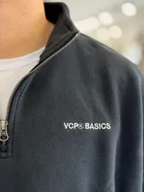 Buzo gris oscuro de corte oversize con cuello alto y cierre frontal parcial. Cuenta con mangas largas y puños elásticos, además de un pequeño bordado con texto en el pecho.