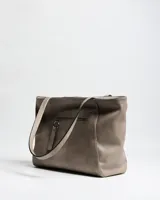 Cartera tipo tote de símil cuero color taupe, con doble asa de hombro, detalles de tachas plateadas en los laterales frontales y placa metálica con logo grabado de la marca XL Extra Large en el centro.