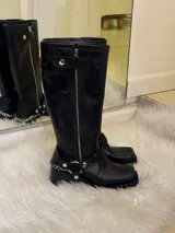 Bota alta tipo biker de cuero negro con cierre metálico lateral que recorre toda la caña. Presenta un diseño versátil con estribo y cadena metálica desmontables en la zona del tobillo, taco cuadrado y plataforma delantera.